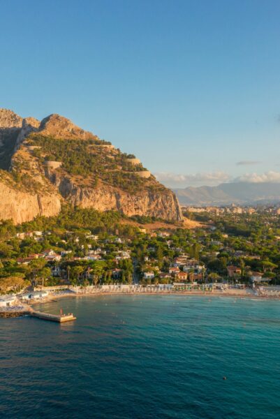 Coastal town at the base of a rocky hill, with a sandy beach and calm blue sea.