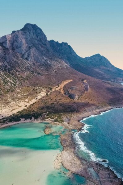 Aerial view of a rugged coastline: turquoise lagoon in a sandy inlet beside rocky mountains and blue sea, under a clear sky.