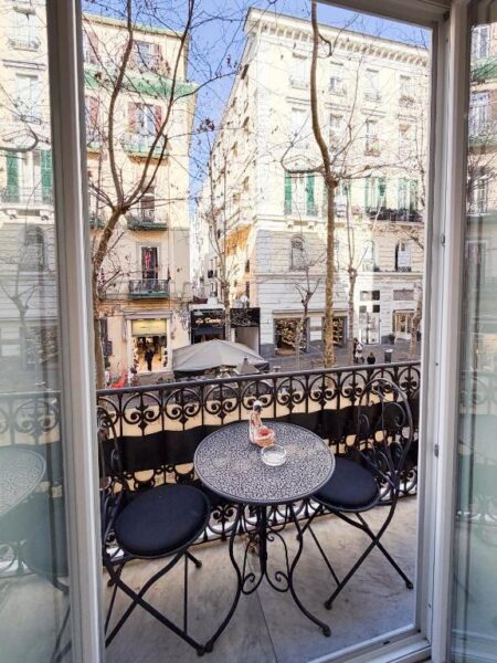 View from a small balcony: round metal table with two matching chairs, ornate railing, overlooking a city street with storefronts and pedestrians.