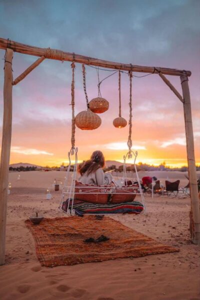 Woman sits on a hanging swing bed beneath woven lanterns in a desert at sunset, with a patterned rug on the sand and warm twilight colors in the sky.