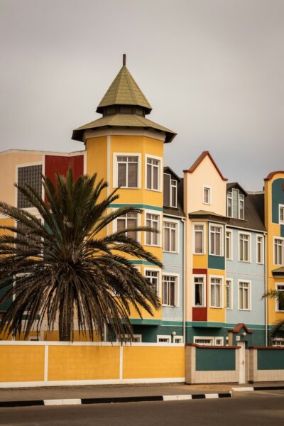 Colorful row of seaside townhouses with a yellow turret, palm tree in the foreground, and a yellow wall along the sidewalk.