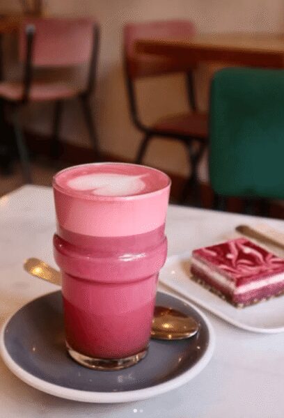 Pink layered latte with a heart-shaped foam on top, served on a saucer beside a pink frosted cake in a cafe.