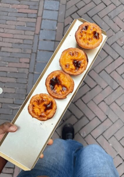 Four custard tarts on a long white serving tray, held by a person in jeans, with a brick-paved sidewalk in the background.
