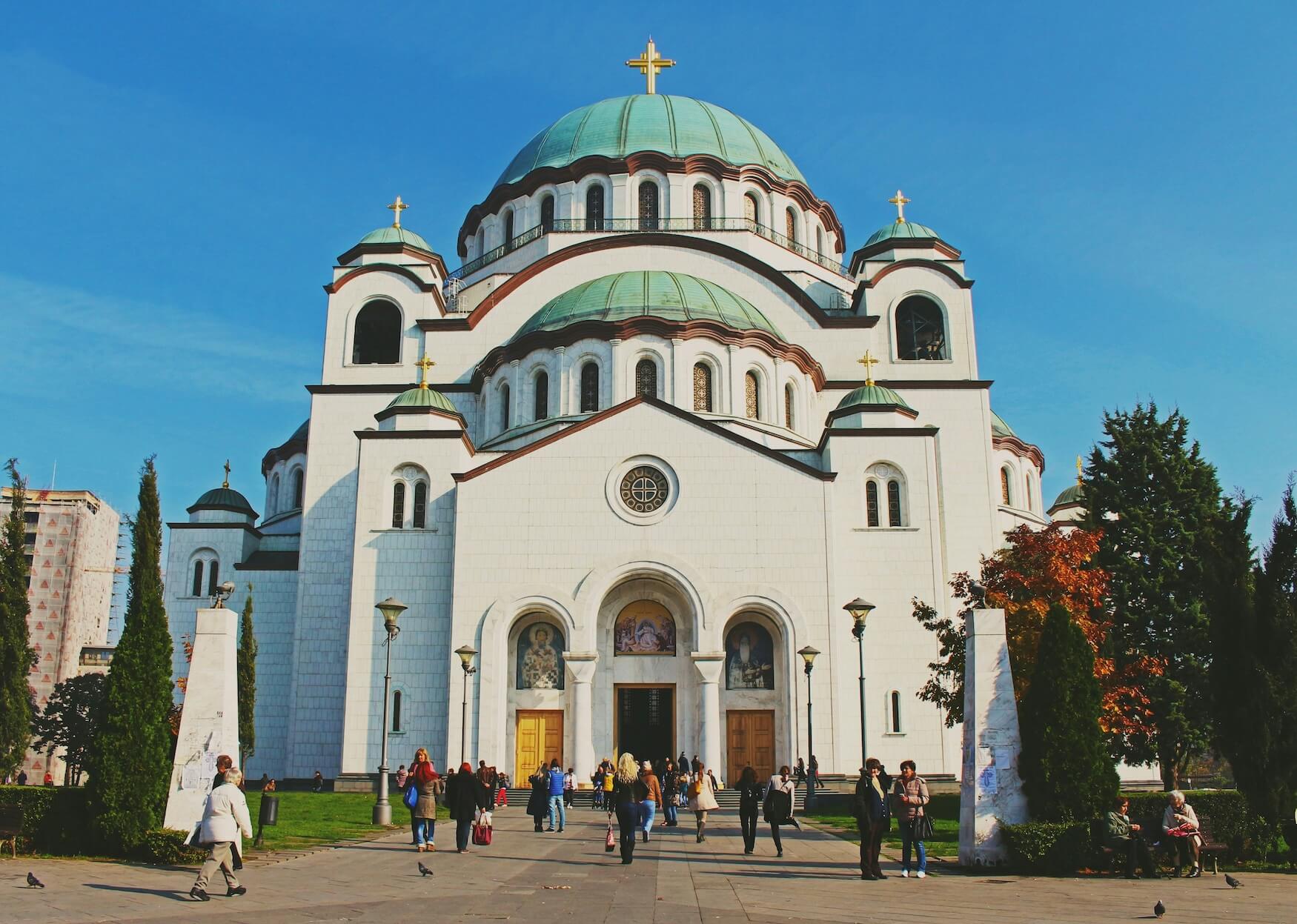 White Orthodox church with green domes and gold crosses under a blue sky, with visitors in the foreground.