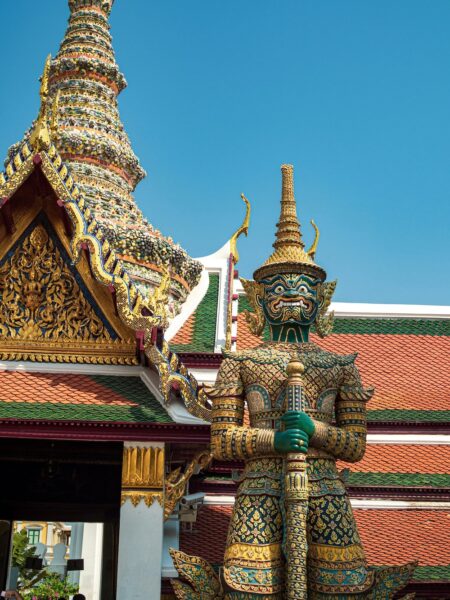 Colorful guardian statue in ornate armor holding a staff in front of a Thai temple with a mosaic roof and blue sky.