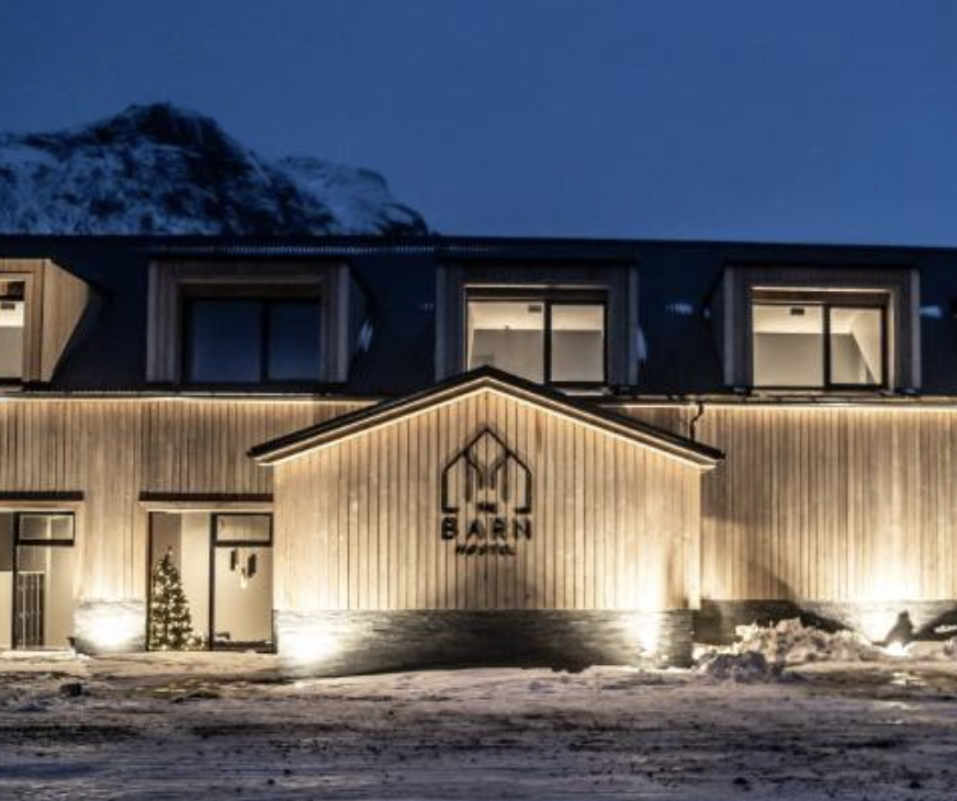 Exterior view of a wooden lodge at dusk, with warm lights outlining the roof and snow on the ground.