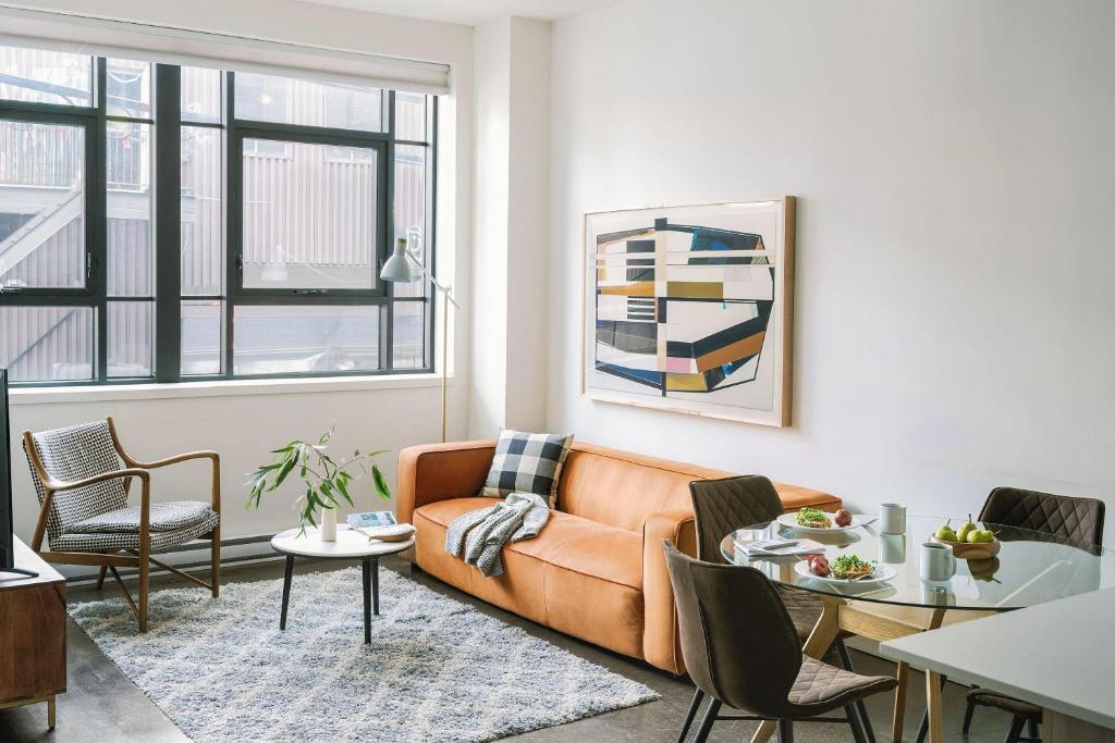 Bright living room with an orange sofa, plaid cushion, and abstract wall art next to a glass dining table set for a light meal.