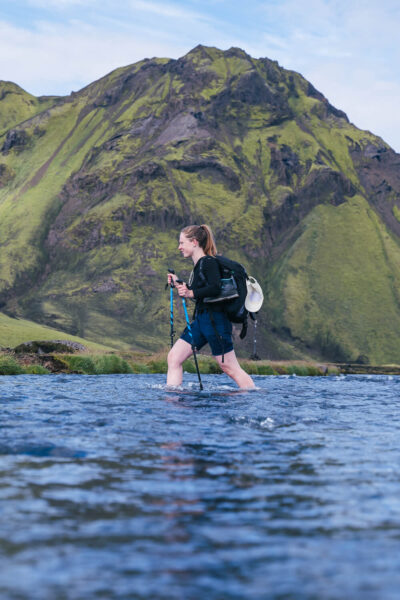 Hiker with a backpack and trekking poles wading through a shallow river, green mountains in the background.