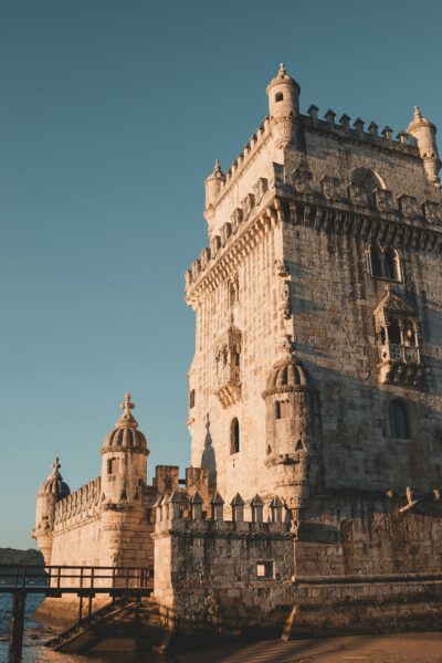 Stone fortress tower by the water with crenellated battlements and decorative turrets bathed in warm sunlight, blue sky above.