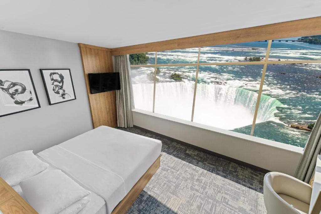 Hotel room with a double bed and white linens, wall-mounted TV, and a large window showing a waterfall outside.