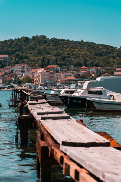 Harbor with moored boats along a weathered wooden pier and a hillside town in the background.