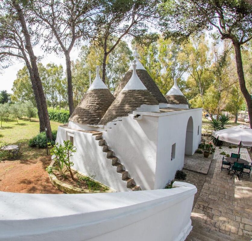 White cone-roofed trulli houses with stone stairs in a sunlit garden setting.