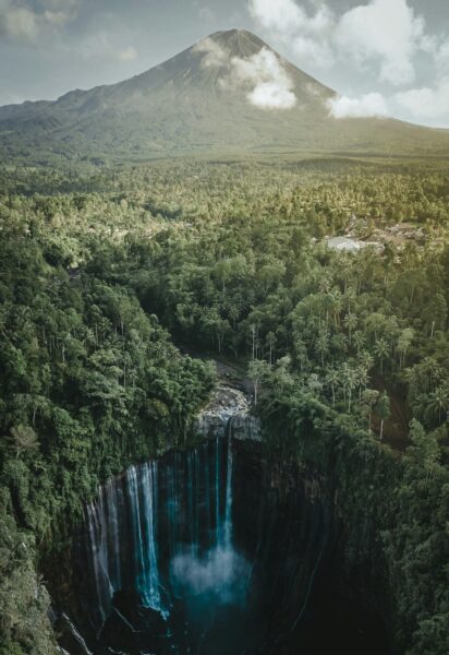 Aerial view of a conical volcano rising above dense tropical forest, with a bright turquoise waterfall cascading into a deep gorge in the foreground
