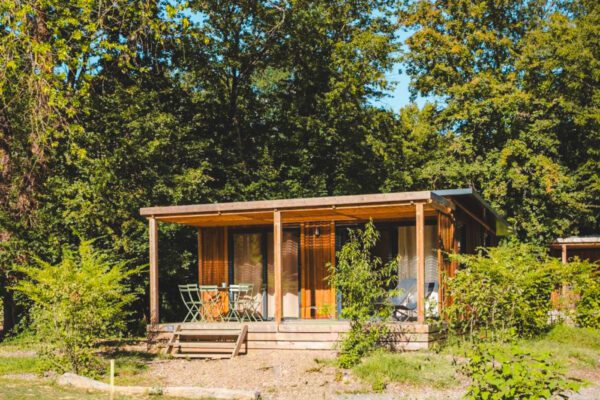 Wooden cabin with glass walls and a patio of chairs, set among trees in a sunny forest clearing