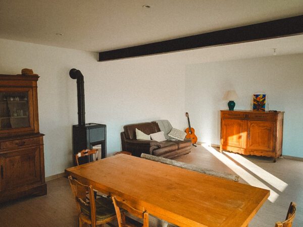 Cozy living room with a wooden dining table in the foreground, brown sofa, guitar, and a wood-burning stove against white walls, sunlit.