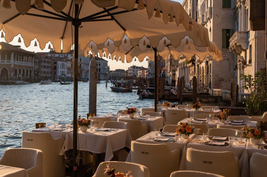 Outdoor waterside restaurant with white-tablecloth tables and floral centerpieces under large beige umbrellas along a canal.