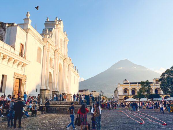 Large white colonial church façade on the left with a crowd in a cobblestone plaza and a mountain in the background under a blue sky
