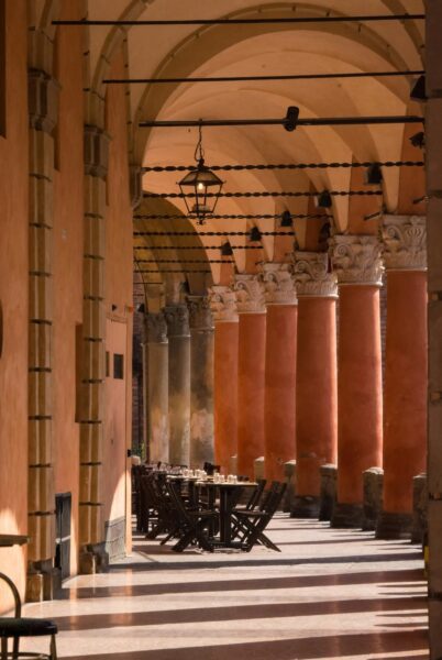Sunlit arcade with tall orange columns, stone capitals, and a row of outdoor dining tables and chairs under arches, lit by a hanging lantern.