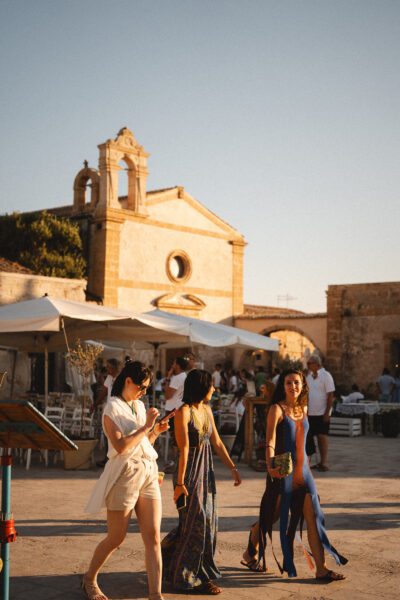 Sunlit plaza with a historic stone church, white tents, and people strolling in summer outfits.