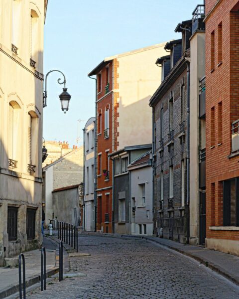 Cobblestone street lined with narrow brick and stucco buildings, black street bollards, and a vintage lamp post under a clear blue sky.