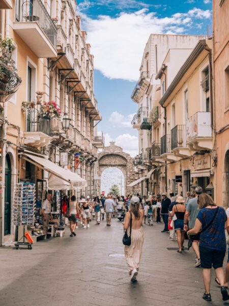 Bustling European street scene with people strolling under warm plaster buildings and an ancient arched opening at the end, blue sky above