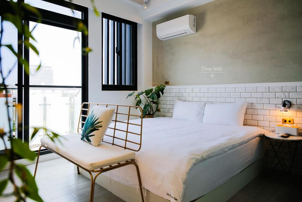 Modern hotel room with a white bed, metal-framed bench at the foot, potted plant, and a balcony door; grey plaster wall and white subway-tile headboard scene.