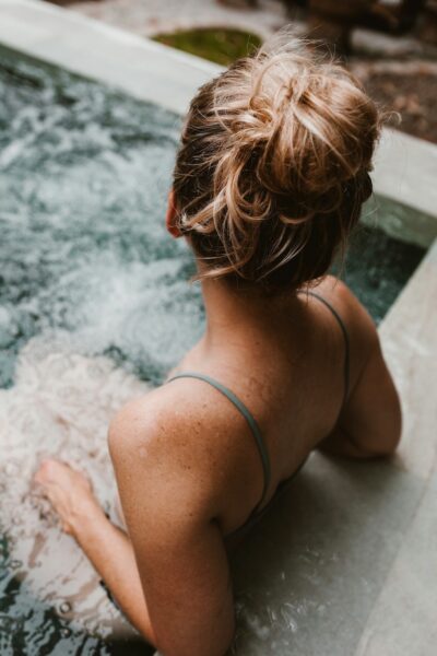 Back view of a person with a messy bun, wearing a teal spaghetti-strap swimsuit, sitting in a bubbling hot tub.