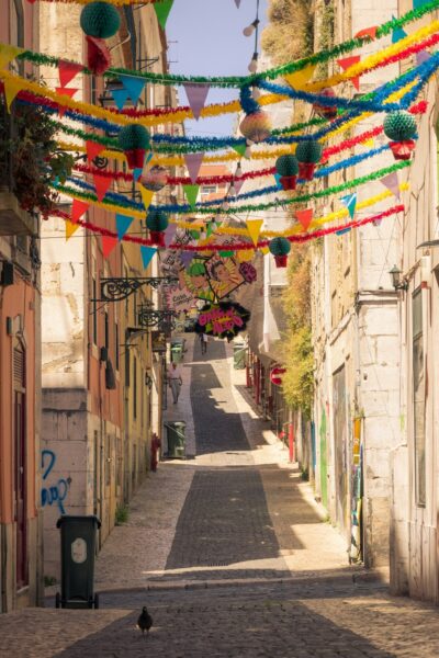 Colorful festival street: pennant banners and paper lanterns across a narrow sunlit cobblestone alley.