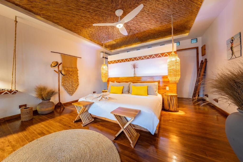 Tropical hotel bedroom with a wooden headboard, white bedding, yellow throw pillows, and woven pendant lights flanking the bed.