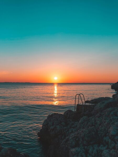 Sunset over the sea with an orange glow on the horizon, rocky shore in the foreground and a metal ladder entering the water.