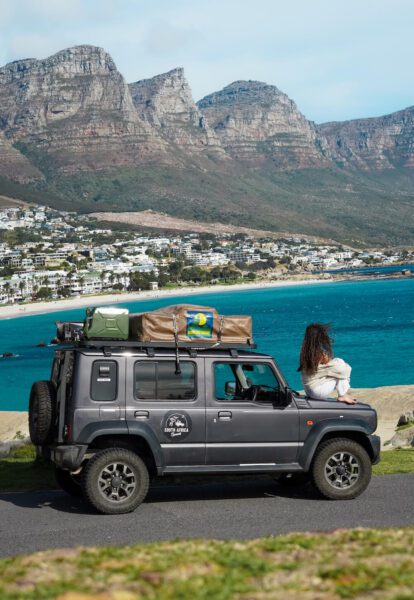 SUV parked by a coastal road with the ocean and town in the background; a person sits on the front fender gazing toward the sea, roof cargo secured on top.