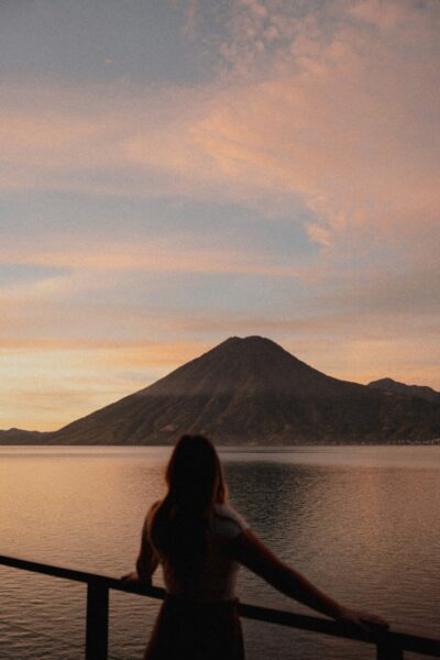 Silhouette of a person standing by a railing, gazing at a lake with a cone-shaped mountain at sunset.