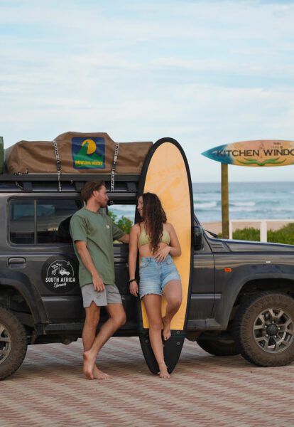 Couple leaning on a dark SUV with a yellow surfboard and rooftop bag labeled 'Howling Moon' beside a beach.